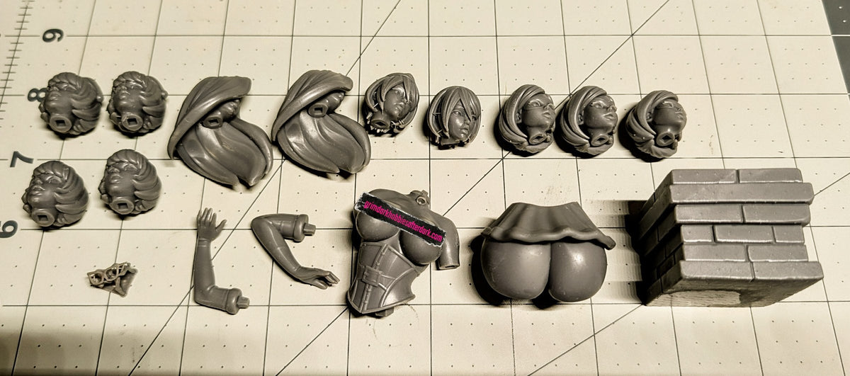 Unpainted grey resin parts from a large pin-up kit laid out on a cutting mat: heads, hair, arms, torso, hips and a brick base